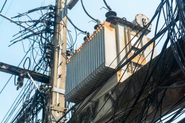 Elektrik direğine monte edilmiş elektrik transformatörünün yakın plan görüntüsü, karmaşık kablolarla çevrili, kentsel elektrik altyapısı ve bakım gereksinimleri gösteriliyor.