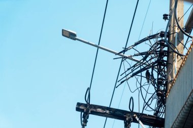 Şehir elektrik hatlarının detaylı görüntüsü ve açık mavi gökyüzüne karşı bir sokak lambası. Sahne modern bir şehirdeki altyapının karmaşıklığını yakalıyor..