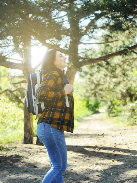 Smiling happy woman in yellow checkered shirt with a traveling bag spends time in the forest. Summer time holidays concept.