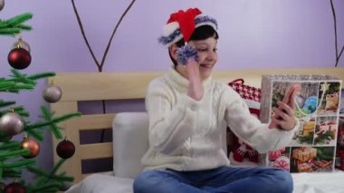 Noel çevrimiçi selamlar. Noel Baba şapkalı gülümseyen çocuk aile ve arkadaşlarıyla video görüşmeleri için cep telefonu kullanıyor. 4K 'da çekilmiş.