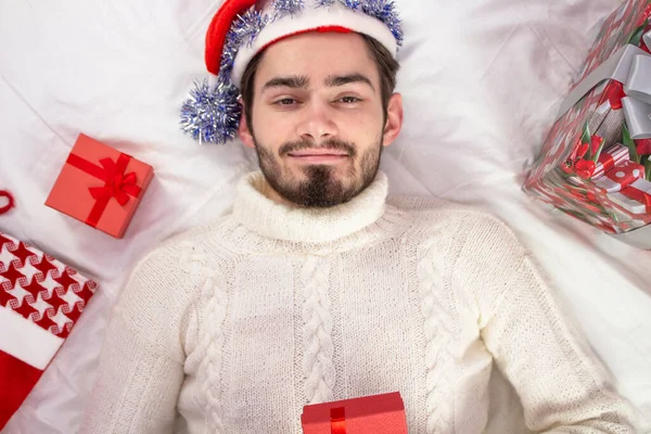 İnternette tebrikler. Noel çevrimiçi selamlar. Sakallı mutlu adam kameraya bakıyor ve gülümsüyor. Büyük bir hediyenin ellerinde. Adam beyaz yatakta yatıyor.