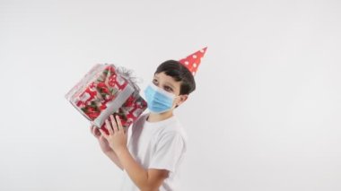 Doğum günü. Hediye. Doğum günü için kırmızı şapkalı ve koruyucu maskeli bir çocuğun portresi. Çocuk bir hediye tutuyor ve baş parmağını kaldırıyor. Karantinaya alın. 4K