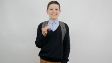 Okul kıyafetli, sırtında sırt çantasıyla neşeli bir çocuk elinde poposuyla beyaz bir arka planda kameraya gülümsüyor. Okula dönelim. 4k