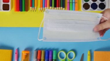 Sarı-mavi arka plandaki okul ofisi boyalar, kalemler, bir makas ve koruyucu bir maske. 4K