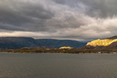 Sarmiento Kanalı, Şili - 11 Aralık 2008: Amalia Buzulu ve Fiyort. Kayalık sahildeki bir parça güneş ışığı gri okyanus suyu ve kara bulutlarla tezat oluşturuyor. Yeşil yapraklı küçük adalar.