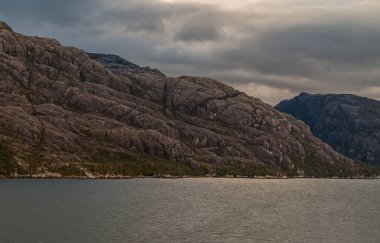 Sarmiento Kanalı, Şili - 11 Aralık 2008: Amalia Buzulu ve Fiyort. Kayalık sahildeki çapraz çizgiler yukarıdaki karanlık bulutlarla ışık yansıtan suya iner. Su kenarında yeşil yapraklar..