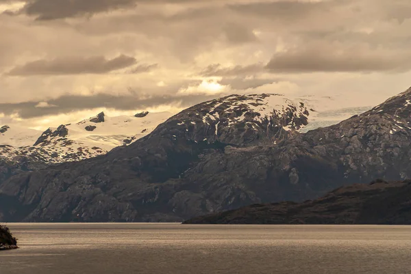 Sarmiento Channel 2008年12月11日 Amalia Glacier Fjord 暗い岩の山の頂上の上に軽いパッチで茶色の雲の下の山の範囲で雪 水辺の緑の葉 — ストック写真