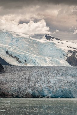 Sarmiento Kanalı, Şili - 11 Aralık 2008: Amalia Buzulu. Mavimsi erime noktasının bir kısmının portresi ve kahverengi yağmurlu bulutların altındaki kayalık dağlarda buzul akıntısını gösteriyor.