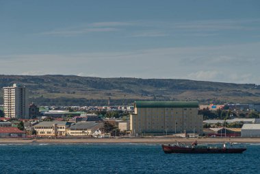 Punta Arenas, Şili - 12 Aralık 2008: Koyu mavi sularda teknesi olan sahil şeridi, uzun ve kısa binalar ve açık mavi bulutlar altında yeşil tepe..