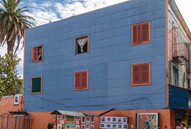 Buenos Aires, Arjantin 19 Aralık 2008, La Boca Mahallesi. Kestane rengi pencereler ve kepenkler, bazıları açık, gökyüzü mavisi ev duvarı. Kenarlarında yeşil yapraklar.