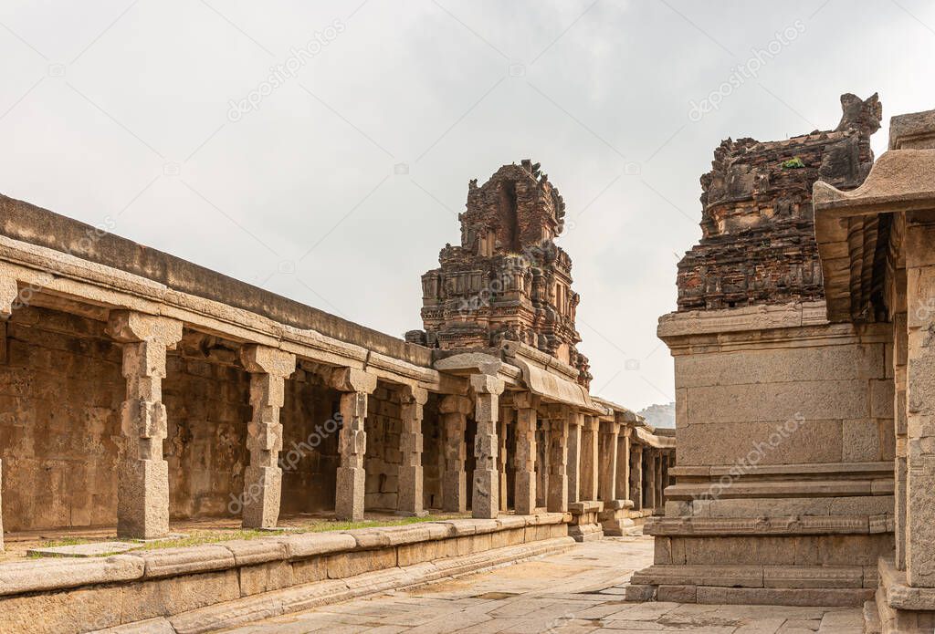 Hampi, Karnataka, India - November 5, 2013: Sri Krishna temple in ruins. Brown stone demolished back of side entrance Gopuram under silver sky and standing on beige stone pillared hall along outside wall.
