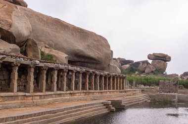 Hampi, Karnataka, Hindistan - 5 Kasım 2013: Sri Krishna tankı harabe halinde. Büyük kahverengi kaya, gümüş gökyüzünün altında havuzun kenarındaki yıkıcı galerinin üzerinde asılı duruyor. Arkadaki yeşil yapraklar..