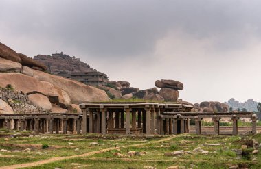Hampi, Karnataka, Hindistan - 5 Kasım 2013: Sri Krishna tankı harabe halinde. Salonun kalıntıları, önünde çimen ve toprak yollar olan gri bulutların altında heykel sütunlar ve galeriler. Her yerde kaya parçaları var..
