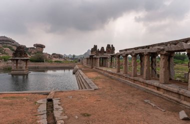 Hampi, Karnataka, Hindistan - 5 Kasım 2013: Sri Krishna tankı harabe halinde. Havuzun güney tarafında, gri bulutların altında tapınak ve galeri var. Ufuktaki kayalar ve dağlar.
