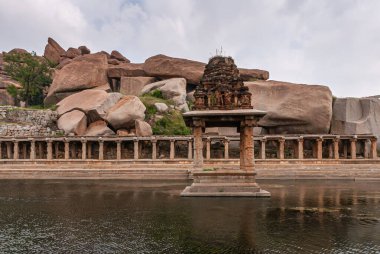 Hampi, Karnataka, Hindistan - 5 Kasım 2013: Sri Krishna tankı harabe halinde. Kahverengi taş hasarlı tapınak, galeri kemerinin önündeki siyah sularda ve arkada mavi bulutların altında büyük balık avcıları..