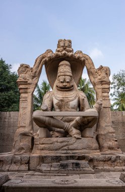 Hampi, Karnataka, Hindistan - 5 Kasım 2013: Lakshmi Narasimha Tapınağı. Mavi gökyüzünün altında, arkasında yeşil yapraklar olan dev kahverengi taş bir tanrı heykeli..