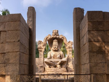 Hampi, Karnataka, Hindistan - 5 Kasım 2013: Lakshmi Narasimha Tapınağı. Mavi gökyüzünün altında, arkasında yeşil yapraklar olan dev kahverengi taş bir tanrı heykeli. Açık hava tapınağının dışından görüldü..