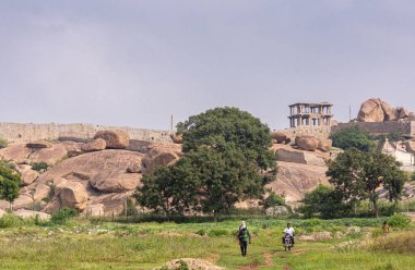 Hampi, Karnataka, Hindistan - 5 Kasım 2013: Badavilinga Tapınağı 'nın dışındaki manzara açık mavi gökyüzü altında koyu kahverengi kaya kuşağı ve önde yeşil bitki örtüsü. Yayalar ve motosikletler.