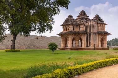 Hampi, Karnataka, Hindistan - 5 Kasım 2013: Zanana Enclosure. Kahverengi taştan Lotus Mahal, yeşil çimenlikte, kırmızı çiçekli ve sarı yapraklı, mavi bulutların altında. Dış duvar arkada..