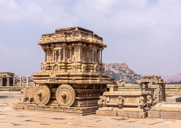 Хампи, Карнатака, Индия - 5 ноября 2013 года: Храм Виджая Виттала. Статуя колесницы из бежевого камня в центре комплекса под голубым небом. Скульптуры и горы на горизонте.