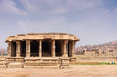 Hampi, Karnataka, Hindistan - 5 Kasım 2013: Vijaya Vitthala Tapınağı. Terk edilmiş ve yıkık edilmiş bir mandapam salonu ve ufukta dağlarla birlikte mavi bulutların altında. Kadın bahçıvanlar renk ekler.