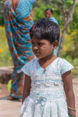 Lakundi, Karnataka, Hindistan - 6 Kasım 2013: Beyaz-mavi elbiseli, kısa siyah saçlı, küpeli ve diğer takılardan oluşan küçük bir kız çocuğu. Sari ve yeşilin solmuş arkaplanı.