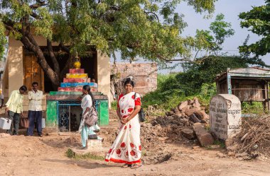 Nandakeshwar, Karnataka, Hindistan - 7 Kasım 2013: Sari 'deki kadınlar ve erkekler yeşil yeşillikler ve açık mavi gökyüzü altında toprak yol boyunca bir halk otobüs durağında bekliyorlar. Renkli sunak.