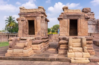 Aihole, Karnataka, Hindistan - 7 Kasım 2013: 2 basit boş türbe kahverengi bina Suryanarayana tapınağının önünde arkasında yeşil yapraklar bulunan mavi bulutların altında. İnsan kıyafetleri renk katar.