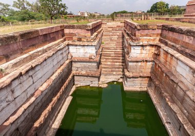 Aihole, Karnataka, Hindistan - 7 Kasım 2013: Huchchimalli Gudi veya Temple. Kahverengi-gri taş duvarlarla çevrili yeşil su tankına inen merdivenlerden merdivenlere bakıyorum..