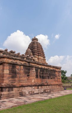 Aihole, Karnataka, Hindistan - 7 Kasım 2013: Huchchimalli Gudi veya Temple. Üzerinde mavi bulutların altında yükselen yontulmuş vimanam ile yan kırmızı taş duvar. Önde yeşil çimen.