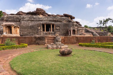 Aihole, Karnataka, Hindistan - 7 Kasım 2013: Ravanaphadi Mağarası Shiva Tapınağı. Mavi bulutların altında kahverengi taş bina eklemeleri olan siyah kaya. Yeşil çimen ve park ön tarafta..