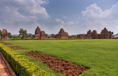 Bagalakote, Karnataka, Hindistan - 7 Kasım 2013: Pattadakal Tapınağı Kompleksi. Mavi bulutların altında kahverengi binalar ve önünde toprak olan yeşil çimenler üzerinde geniş bir manzara..