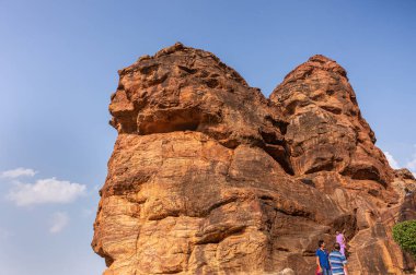 Badami, Karnataka, Hindistan - 7 Kasım 2013: Agasthya Gölü üzerindeki mağara tapınakları. Mavi bulutların altındaki tapınaklara giden dar adımlı kırmızı uçurumlar. İnsan kıyafetleri renk katar..