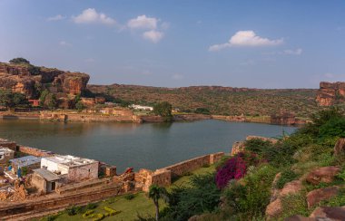 Badami, Karnataka, Hindistan - 7 Kasım 2013: Mavi bulutların altındaki tepeler, kayalar ve kayalıklarla çevrili Agasthya Gölü üzerindeki mağara tapınakları. Çiçekler renk ekler, yeşil yapraklar.