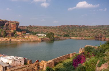 Badami, Karnataka, Hindistan - 7 Kasım 2013: NE-view 'de Agasthya Gölü üzerindeki mağara tapınakları, mavi bulutların altındaki tepeler, kayalar ve kayalıklarla çevrili. Çiçekler renk ekler, yeşil yapraklar.