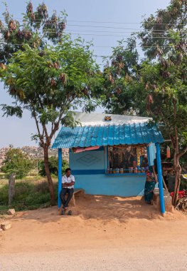 Ballapur, Karnataka, Hindistan - 9 Kasım 2013: Mavi gökyüzünün altındaki kahverengi kumlara kurulmuş, yeşil yaprakların altındaki küçük, mavi, kuru erzak tezgahının dışındaki tedarikçi.