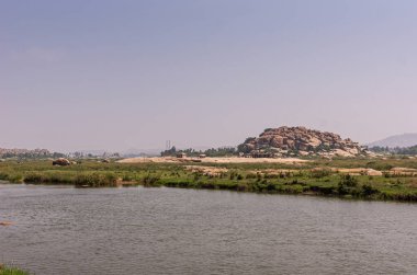 Anegundi, Karnataka, Hindistan - 9 Kasım 2013: Gray Tungabhadra Nehri. Açık mavi gökyüzünün altındaki yeşil adasında Sooryanarayana Tapınağı kompleksiyle geniş bir manzara. Arkadaki kahverengi kayalık tepe.