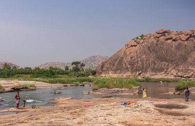 Anegundi, Karnataka, Hindistan - 9 Kasım 2013: Navabrindavana Adası ve Tapınağı. Tungabhadra Nehri 'nde uzun kahverengi kaya tepeleri ve yeşil ağaçların arasında yıkanan insanlar. Giysi renk katar.