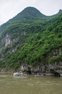 Guilin, Çin - 10 Mayıs 2010: Li Nehri boyunca. Koyu kayalıklardaki yeşil su seviyesindeki mağaralar kısmen yeşil ağaçlarla kaplı gümüş gökyüzünün altındaki yüksek dağlar..