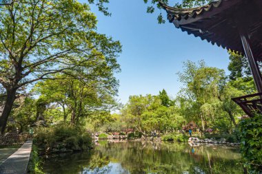 Suzhou, Çin - 3 Mayıs 2010: Mütevazı Yöneticiler Bahçesi. Mavi gökyüzünün altındaki yeşil yapraklar arasında yansıyan göletin geniş manzarası. Çin Mimarlık binasının köşesinde. Çiçekler renk ekler.