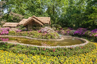 Suzhou, Çin - 3 Mayıs 2010: Mütevazı Yöneticiler Bahçesi. Pembe ve sarı çiçeklerin arasında küçük bir göletin arkasında bej samandan bir çatı çardağı. Yeşil yapraklarla çevrili..