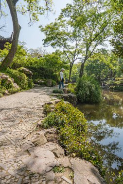 Suzhou, Çin - 3 Mayıs 2010: Mütevazı Yöneticiler Bahçesi. Kahverengi su deresi boyunca gri taş patika ve rüya gören bir kadın. Açık mavi gökyüzünün altında yeşil yapraklar. 