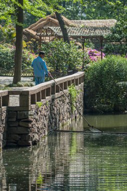 Suzhou, Çin - 3 Mayıs 2010: Mütevazı Yöneticiler Bahçesi. Mavi elbiseli işçiler yeşil su deresinden çöp topluyor. Gri taş duvarın üzerinde yeşil yapraklar ve pembe çiçeklerle duruyorlar..