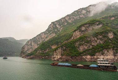 Yangtze Nehri, Three Gorges, Çin - 6 Mayıs 2010: Zigui bölgesi. Yeşil ormanlık tepelerin ve dağların önünde sisli gökyüzünün altında kıyı şeridinde beig-kahverengi kayalıkları olan iki mavna yeşil sularda yol alır..