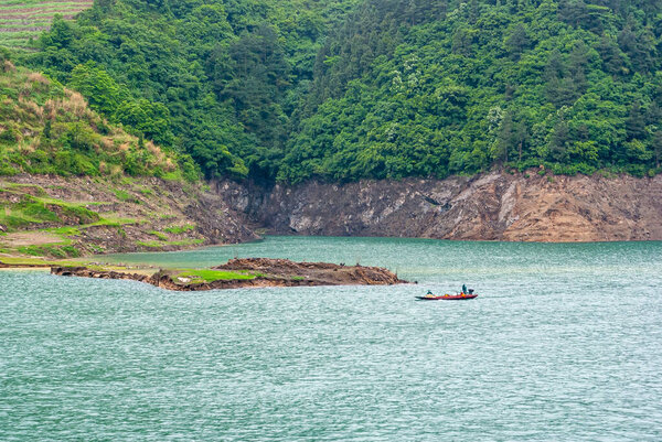 Река Янцзы, Три ущелья, Китай - 6 мая 2010 года: район Сянсичунь в ущелье Силин. Небольшой моторизованный шлюп на зеленой воде перед лесной береговой линией и террасой сельского хозяйства.