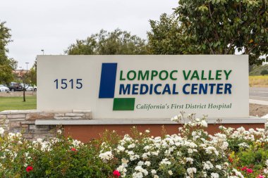 Lompoc, CA, USA - 26 Mayıs 2021: Lompoc Valley Tıp Merkezi 'nin girişinde mavi üzerine beyaz amblem. Otomobiller ve bitkiler gri gökyüzünün altına renk katar.