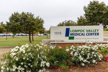 Lompoc, CA, USA - 26 Mayıs 2021: Lompoc Valley Tıp Merkezi 'nin girişindeki sokak numarasının üzerindeki mavi amblemde beyaz. Otomobiller ve bitkiler gri gökyüzünün altına renk katar.