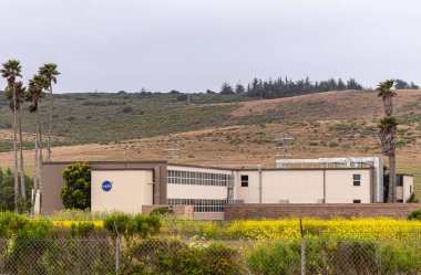Lompoc, CA, USA - 26 Mayıs 2021: Vandenberg Uzay Üssü. Yeşil hardal çiçekleri ve açık mavi gökyüzünün altında yeşil hardal çiçekleri eşliğinde tepelik yeşil manzaralı Nasa binasına yakın çekim. 