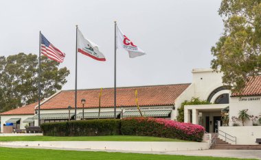 Santa Barbara, CA, USA - 2 Haziran 2021: Şehir kolej tesisleri. ABD, Kaliforniya ve SBCC bayrakları ve Luria Kütüphanesi binası yeşil çimlerin ve gümüş gökyüzünün altında kırmızı çiçekler.
