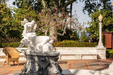 San Simeon, CA, USA - 12 Şubat 2014: Hearst Castle. Ana girişteki fıskiyenin önünde beyaz çıplak heykel arkada yeşil yapraklar, açık mavi gökyüzünün altında kırmızı çiçekler..